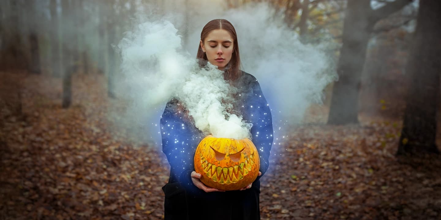 Jeune femme tenant une citrouille sculptée dégageant de la fumée magique, symbole d’Halloween et de sa dimension spirituelle liée à Samhain et au voile entre les mondes.